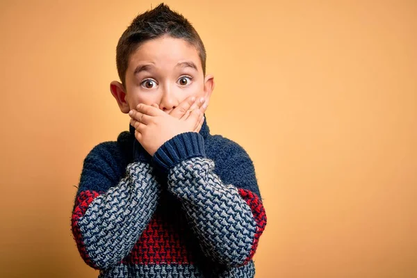 黄色の孤立した背景に冬のセーターを着ている若い男の子は間違いのために手で口をカバーショックを受けました 秘密概念 — ストック写真