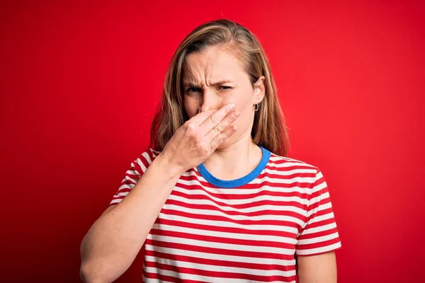 若い美しいブロンドの女性は 孤立した赤い背景の上にカジュアルな縞模様のTシャツを着て 鼻の上に指で息を保持し 臭いや嫌な 耐え難い匂いを嗅いでいます 匂いが悪い — ストック写真