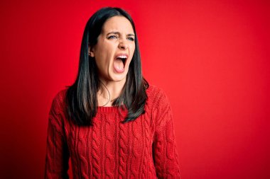 Mavi gözlü genç esmer kadın izole edilmiş kırmızı arka plan üzerine gündelik süveter giyiyor kızgın ve öfkeli çığlıklar atıyor, öfkeyle bağırıyor. Öfke ve agresif kavram.
