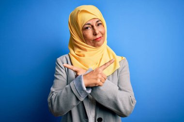 Orta yaşlı esmer iş kadını. Mavi arka plan üzerine geleneksel başörtüsü takıyor. Her iki tarafı da parmaklarıyla işaret ediyor.