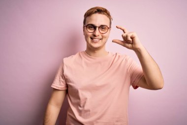 Genç, kızıl saçlı, günlük tişört giyen, pembe arka planda duran, gülümseyen, kendine güvenen, el hareketi yapan, parmaklarıyla işaret yapan ve kameraya bakan genç bir adam. Ölçü kavramı.