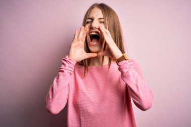 Genç, güzel, kızıl saçlı bir kadın pembe arka plan üzerine günlük süveter giyip elleri ağzında kızgın bir şekilde bağırıyor.