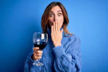 Genç, güzel, esmer bir kadın, izole edilmiş mavi arka planda bir bardak kırmızı şarap içiyor. Ağzı kapalı, utancından ağzı açık, korkusunun dışa vurumu, sessizlikten korkması, gizli konsept.