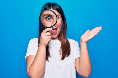 Genç, güzel esmer kadın, izole edilmiş mavi arka plan üzerinde büyüteç kullanıyor başarıyı mutlu bir gülümsemeyle ve kazanan ifadeyle el kaldırarak kutluyor.