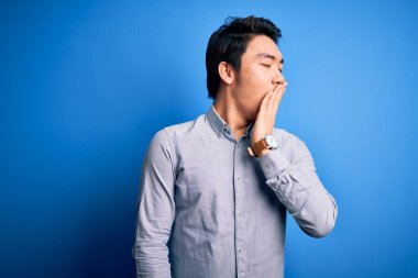 İzole edilmiş mavi arka planda dikilen rahat gömlekli genç, yakışıklı Çinli adam ağzıyla esnemekten sıkılmış. Huzursuz ve uykulu..