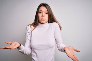 İzole edilmiş beyaz arka plan üzerinde duran sıradan bir kazak giyen genç ve güzel esmer kız elleri ve kolları havada, kafası karışmış bir ifade takınıyor. Şüphe kavramı.