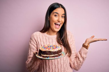 Genç, güzel esmer kadın pembe arka planda doğum günü pastasını tutuyor. Çok mutlu ve heyecanlı, kazanan yüz ifadesi zaferi büyük bir gülümsemeyle kutluyor ve ellerini kaldırıyor.