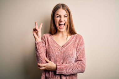 İzole edilmiş beyaz arka planda pembe kazak giyen güzel, kızıl saçlı kadın kameraya gülümseyip parmaklarıyla zafer işareti yapıyor. İki numara..