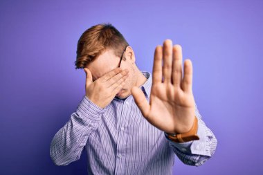 Genç ve yakışıklı kızıl saçlı bir adam mor arka plan üzerine günlük gömlek ve gözlük takıyor gözleri ellerle örtüyor ve üzgün ve korku ifadeleriyle dur hareketi yapıyor. Utanç verici ve olumsuz bir kavram..