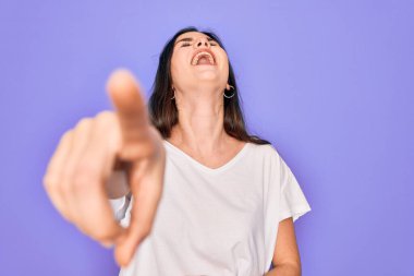 Genç, güzel esmer kadın, mor arka plan üzerine günlük beyaz tişört giyip sana gülüyor, eli vücudunun üzerinde kamerayı işaret ediyor, utanç ifadesi takınıyor.