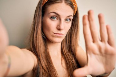 Diadem giyen genç, güzel, kızıl saçlı bir kadın kameranın önünde dur işareti yaparken ciddi ve kendinden emin bir ifadeyle, savunma jesti yapıyor.