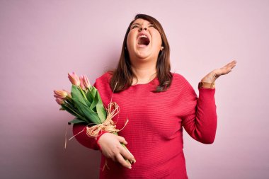 Güzel, iri yarı bir kadın pembe arka planda romantik lale çiçekleri tutuyor deli deli deli bağırıyor ve agresif bir ifadeyle bağırıyor ve kollarını kaldırıyor. Hayal kırıklığı konsepti.