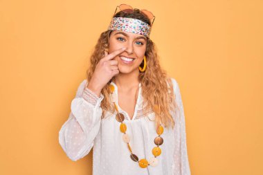 Mavi gözlü, güneş gözlüğü takan ve aksesuar takan el ve burnu işaret eden, neşeli gülümseyen güzel sarışın hippi kadın. Güzellik konsepti