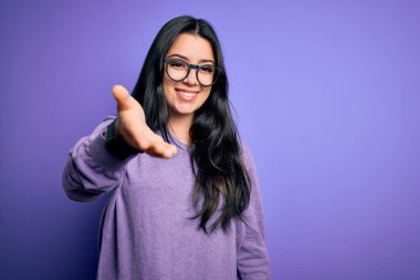 Mor izole edilmiş arka planda gözlük takan esmer bir kadın dostça bir selamlama ve hoş geldin hediyesi olarak el sıkışmayı sunuyor. Başarılı bir iş..