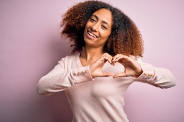 Afro saçlı, pembe arka planda sıradan bir süveter giyen, elleri ile kalp sembolü şeklinde gülümseyen Afro-Amerikalı genç bir kadın. Romantik konsept.
