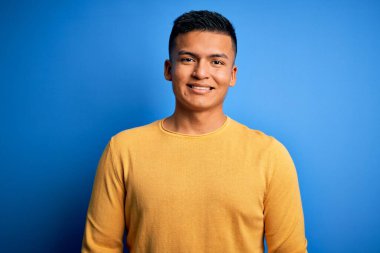 İzole edilmiş mavi arka plan üzerinde sarı günlük süveter giyen yakışıklı Latin bir adam. Yüzünde mutlu ve havalı bir gülümseme var. Şanslı kişi..