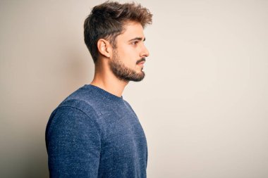 Sakallı, sıradan bir süveter giyen, beyaz arka plan üzerinde duran, rahat bir görünüm takınan, kendine güvenen gülümseyen genç yakışıklı bir adam..