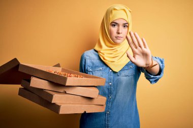 Müslüman başörtüsü takan genç bir kız sarı arka planda İtalyan pizzasıyla kutuları tutarken dur işareti yaparken ciddi ve kendinden emin bir ifadeyle savunma hareketi yapıyor.