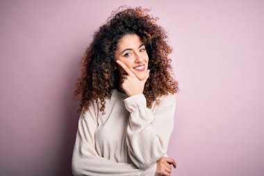 Kıvırcık saçlı ve piercingli güzel bir kadın pembe arka planda rahat bir süveter giyiyor kameraya bakıyor kolları çapraz ve çenesiyle gülümsüyor. Olumlu düşünüyorum..