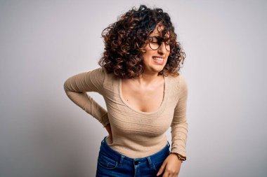 Genç, kıvırcık, kıvırcık, rahat tişört giyen, beyaz arka planda gözlük takan sırt ağrısından muzdarip bir kadın.