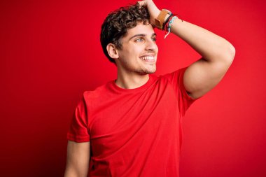 Kıvırcık saçlı, kırmızı arka planda rahat tişörtler giyen, el hareketleriyle saçlarına dokunan, çekici ve modaya uygun pozlar veren genç, sarışın, yakışıklı bir adam.