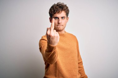 Young blond handsome man with curly hair wearing casual sweater over white background Showing middle finger, impolite and rude fuck off expression