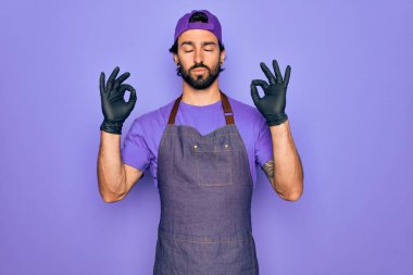 Profesyonel önlük giyen yakışıklı İspanyol bir adam dövme sanatçısı olarak çalışıyor gözleri kapalı gülümsüyor parmaklarıyla meditasyon hareketi yapıyor. Yoga konsepti.