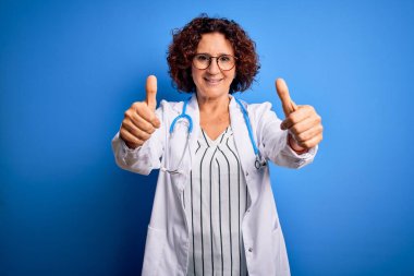 Mavi arka plan üzerinde steteskop ve ceket giyen orta yaşlı kıvırcık saçlı kadın el hareketini onaylıyor, gülümsüyor ve başarı için seviniyor. Kazanan jest.
