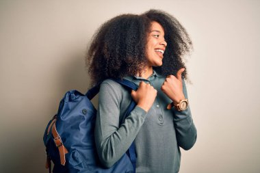 Afro saçlı, arka planda sırt çantası takan ve gülümseyen yüzüyle baş parmağıyla yüzünü gösteren Afro Amerikalı genç bir kız öğrenci.