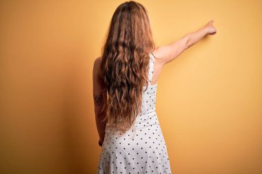 Genç, sarışın, tatilde. Sarı arka planda elbisesi var. Önünü işaret ediyor.