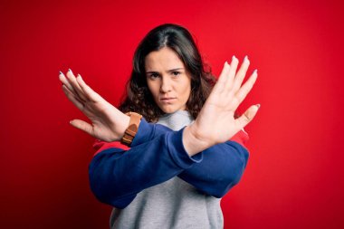 Kıvırcık saçlı genç bir kadın izole edilmiş kırmızı arkaplan üzerinde sıradan bir kazak giyiyor. Reddedilmiş yüz ifadesi kollarını kavuşturarak negatif işaret veriyor, kızgın yüz ifadesi takınıyor.