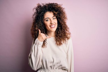 Kıvırcık saçlı ve piercingli güzel bir kadın pembe arka planda rahat bir süveter giyiyor gülümsüyor telefonla el ve parmaklarıyla telefon hareketi yapıyor telefonda konuşuyor gibi. İletişim kavramları.