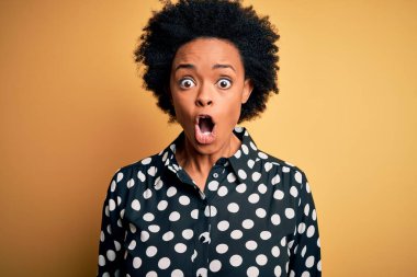 Kıvırcık saçlı, genç, Afro-Amerikan bir kadın. Günlük gömleğini giymiş. Korkmuş ve şok olmuş bir halde duruyor..