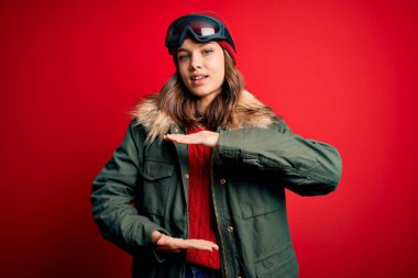 Kayak gözlüğü ve kış havası için kayak gözlüğü takan genç sarışın kız kırmızı zemin üzerinde elleri büyük ve büyük beden işareti, ölçü sembolü göstererek el kol hareketi yapıyor. Kameraya bakıp gülümsüyor. Ölçüm konsepti.