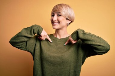 Modern kısa saçlı, sarı arka planda rahat süveter giyen, kendine güler yüzlü, kendini gururlu ve mutlu gösteren genç sarışın kadın..