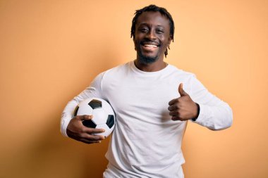 Genç Afrikalı Amerikalı oyuncu futbol topunu sarı arka planda tutarak oynuyor. Kocaman gülümsüyor ve gülümsüyor. İşaret veriyor.