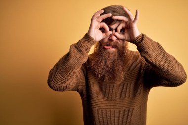 Sarı arka planda sakallı ve kışlık süveter giyen yakışıklı İrlandalı kızıl adam. Dürbün gibi jest yapıyor, gözleri parmaklarından bakıyor. Çılgın ifade.