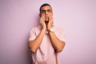 Yakışıklı Afro-Amerikalı adam pembe arka plan üzerinde günlük tişört ve gözlük takıyor. Yüzü, depresyonu ve üzüntüyü örten eller, sorun yüzünden üzgün ve sinirli.