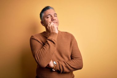 Orta yaşlı, yakışıklı, gri saçlı, sarı arka planda rahat bir süveter giyen ve kameraya kollarını kavuşturup gülümseyerek bakan bir adam. Olumlu düşünüyorum..