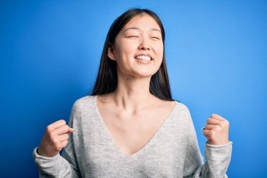 Mavi izole edilmiş arka plan üzerinde sıradan bir süveter giyen genç, güzel Asyalı bir kadın mutlu ve heyecanlı bir şekilde kollarını kaldırıp, gülümseyerek ve başarı için çığlık atarak zafer jesti yapıyor. Kutlama konsepti.