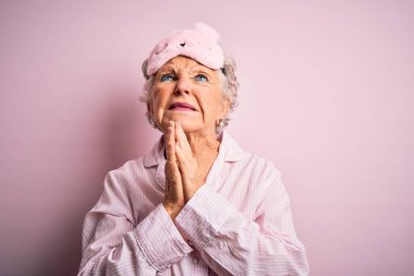 İzole edilmiş pembe arka plan üzerinde uyku maskesi ve pijama giyen son sınıf güzeli bir kadın. Yüzünde umut ifadesi olan ve endişeli bir şekilde el ele yalvarıyor ve dua ediyor. Yalvarıyorum..