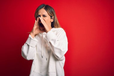 Orta yaşlı güzel sporcu kadın izole edilmiş kırmızı arka plan üzerinde sıradan bir kazak giyiyor elleri ağzında kızgın bir şekilde bağırıyor.