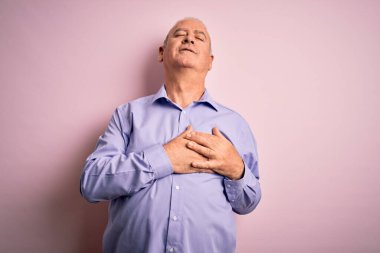 Orta yaşlı yakışıklı, beyaz gömlekli, pembe arka planda dikilen, elleri göğsünde, gözleri kapalı ve yüzünde minnet ifadesi olan bir adam. Sağlık kavramı.