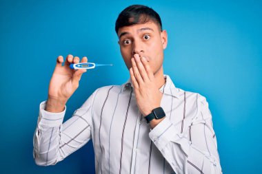 Tıbbi termometreyi mavi izole edilmiş arka plan üzerinde tutan genç İspanyol adam. Ağzı, hata yüzünden utançla kaplanmış, korku ifadesi, sessizlikten korkmak, gizli konsept.
