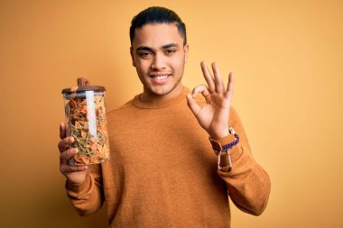 İzole edilmiş sarı arka planda İtalyan makarnası ile dolu kavanozu tutan genç Brezilyalı adam tamam işareti yapıyor, mükemmel bir sembol.