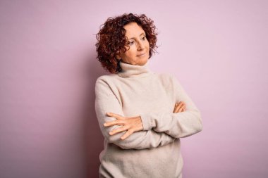 Orta yaşlı güzel kıvırcık saçlı kadın pembe arka planda balıkçı yaka kazak giyiyor. Kollarını kavuşturmuş, ikna olmuş ve kendinden emin bir şekilde yan tarafa bakıyor.