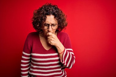 Orta yaşlı güzel kıvırcık saçlı kadın kırmızı arka planda sıradan çizgili süveter giyiyor kendini iyi hissetmiyor ve soğuk algınlığı veya bronşitin belirtisi olarak öksürüyor. Sağlık hizmeti kavramı.