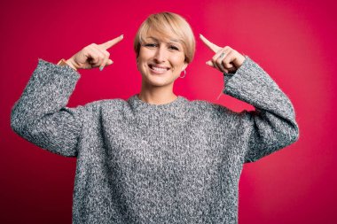 Modern kısa saçlı, pembe arka planda rahat süveter giyen genç sarışın kadın iki eliyle başını gösteren gülümseme, harika bir fikir ya da düşünce, iyi bir hafıza