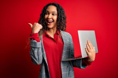 Kıvırcık saçlı genç bir iş kadını kırmızı arka planda dizüstü bilgisayar kullanıyor ve gülümseyen bir gülümsemeyle baş parmağıyla yüzünü gösteriyor.