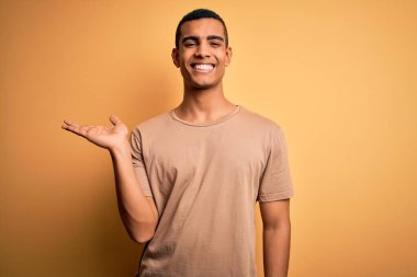 Genç, yakışıklı, Afro-Amerikalı sıradan tişörtler giyen, sarı arka planda dikilen, gülümseyen, elini kaldırıp kameraya bakan bir adam..